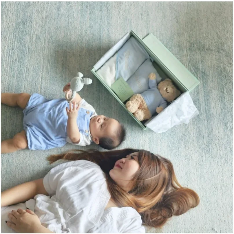 家長和嬰兒躺在打開的禮盒旁邊，盒裡有衣服和毛公仔